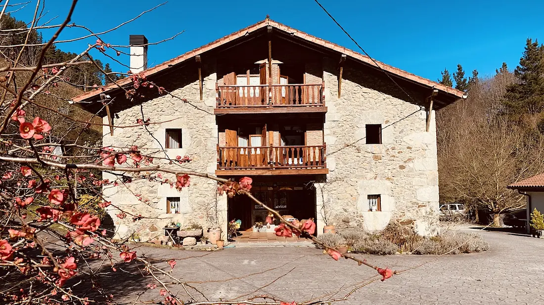 El caserío de Urresti: fachada de piedra con balcón de madera y geranios rojos, rodeado de verde.