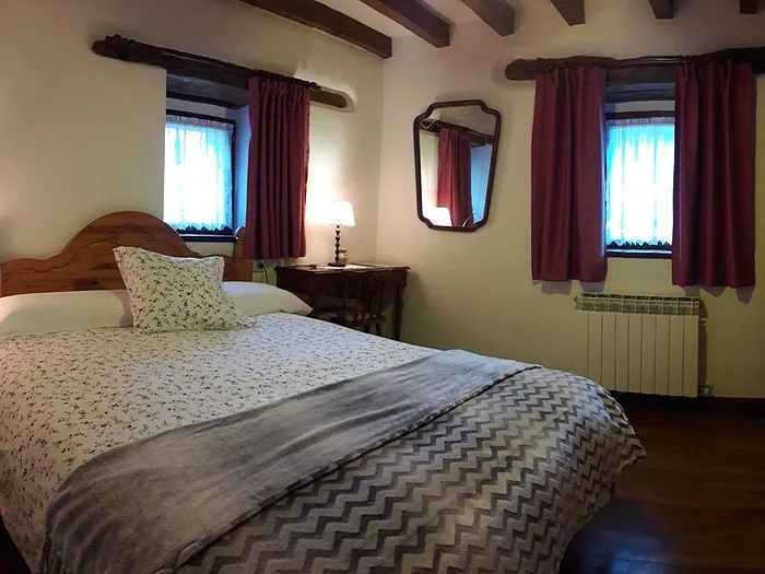 Habitación con dos ventanas pequeñas, cama vestida y mesillas con lámparas encendidas.
